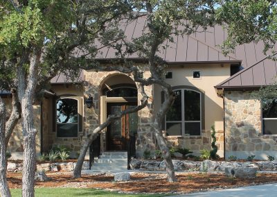 Custom Home Builder. Tan stucco home with brown standing seam metal roof on heavily wooded lot